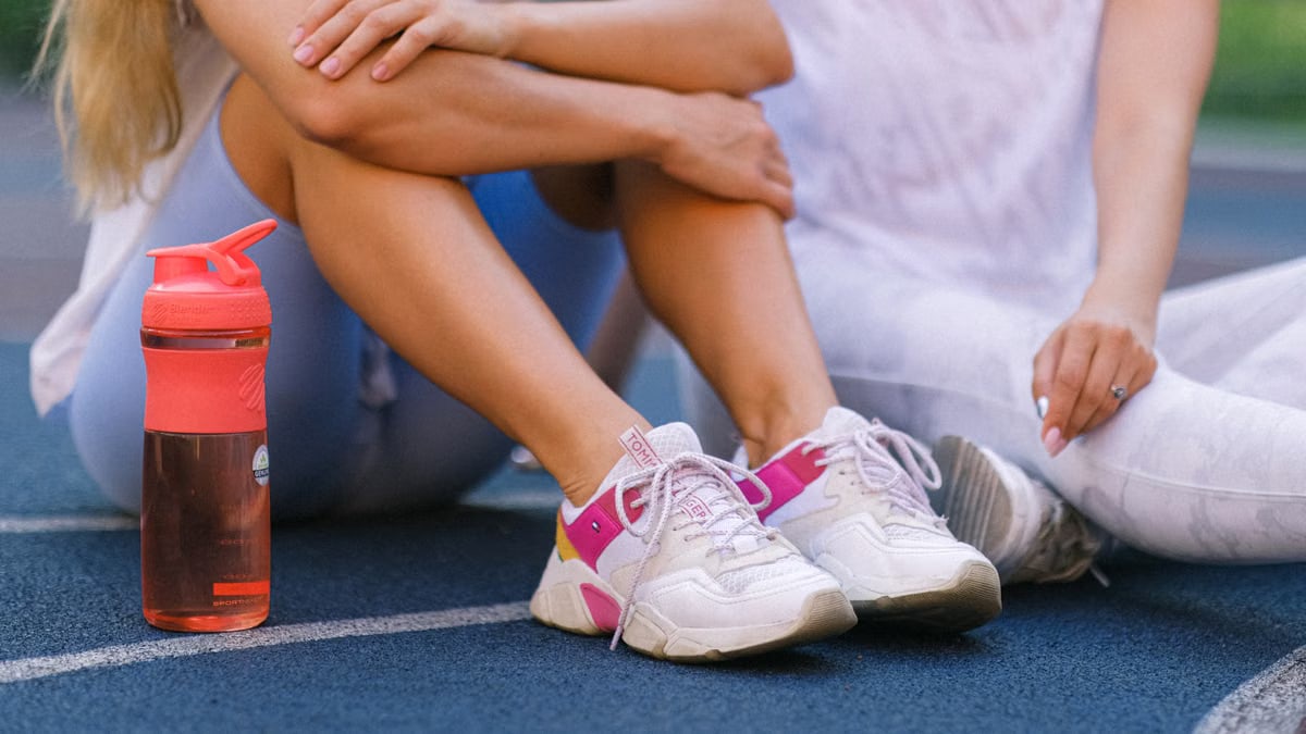 Crop anonymous female friends in activewear sitting on sports ground after training near bottle with water in daytime